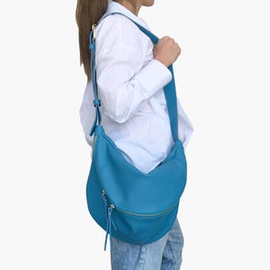 Könnte beinhalten: Eine hellblaue Lederschultertasche mit einem langen Riemen. Die Tasche hat eine geschwungene Form und einen Reißverschluss.