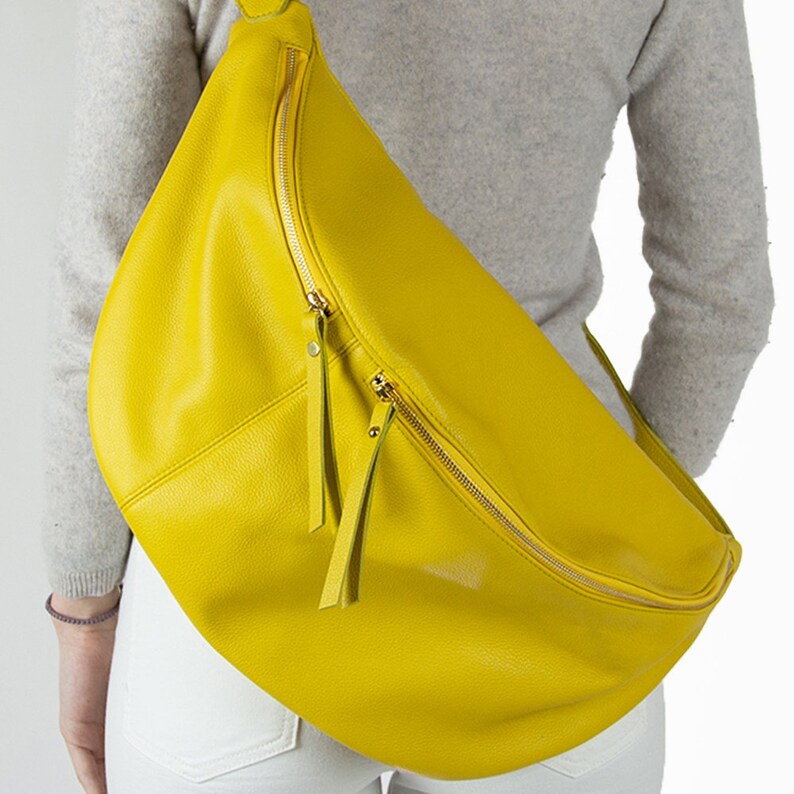 May include: A bright yellow leather crossbody bag with a gold zipper and a long strap. The bag is worn over the shoulder of a person wearing a grey sweater and white pants.