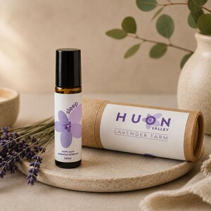 Könnte beinhalten: Eine 10ml Braunglas-Roll-on-Flasche mit schwarzem Verschluss und weißem Etikett, das eine violette Blume und den Text sleep 100% pure essential oil blend zeigt, neben einer passenden zylindrischen Papphülse mit der Aufschrift Huon Valley Lavender Farm.