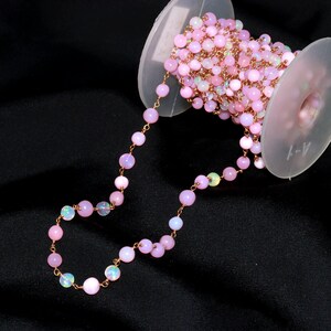May include: A spool of rose gold wire chain featuring round, light pink opal beads spaced evenly along the length. The chain is wound around a translucent plastic spool, with a portion of the beaded chain extending outward in a curved line.