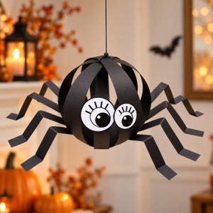May include: A hanging black paper spider decoration featuring a spherical body constructed from intersecting paper strips and two large white circular eyes with black pupils and eyelashes, attached to eight angular, jointed black paper legs.