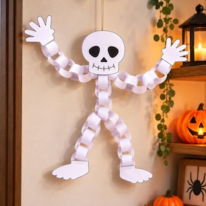Könnte beinhalten: Eine hängende Skelettdekoration aus weißen Papierkettengliedern. Die Figur besteht aus einem flachen Papierschädel mit schwarzen Augenhöhlen, einer Nase und einem aufgemalten Mund. Arme und Beine sind aus ineinandergreifenden Papierschlaufen gefertigt und enden in flachen Papierhänden und -füßen. Eine dünne Schnur ist oben am Schädel zur Aufhängung befestigt.
