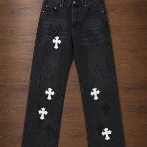 Könnte beinhalten: Schwarze Jeans aus Denim mit geradem Bein, Knopfleiste und fünf Taschen. Der Stoff ist mit mehreren weißen Kreuz-Aufnähern und tonalen, schwarzen Stickereien in Kreuzform verziert, ergänzt durch dekorative Steppnähte oberhalb der Knie.