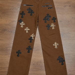 Könnte beinhalten: Braune Hose mit einer Reihe von schwarzen kreuzförmigen Aufnähern und kreuzförmigen Aufnähern mit Leopardenmuster, die über die Beine verteilt sind. Die Hose hat zwei Gesäßtaschen mit schwarzem Besatz und ein kleines rechteckiges Etikett.