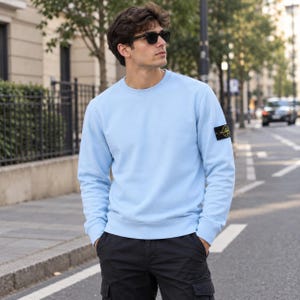 Könnte beinhalten: Ein hellblaues Langarm-Sweatshirt mit Rundhalsausschnitt und einem schwarzen, gelben runden Logo-Aufnäher am linken Ärmel. Das Kleidungsstück hat einen gerippten Ausschnitt, Bündchen und Saum, was diesem lässigen Pullover eine klare Passform verleiht.
