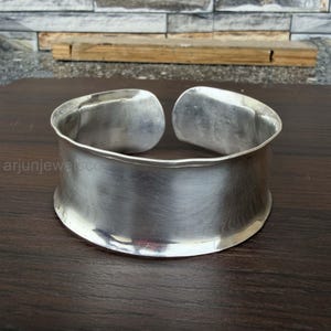 May include: A wide, open-ended silver cuff bracelet with a smooth, polished finish and a slightly curved, concave profile. The metal surface reflects light, and the text arjunjewelsco is visible on the left side of the bracelet.