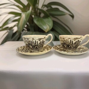Puede incluir: Dos tazas de té de cerámica color crema con sus platos a juego, que presentan un detallado estampado en marrón oscuro de una escena de carruaje tirado por caballos con árboles y elementos paisajísticos. Cada taza tiene un asa curva y borde festoneado.