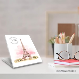 May include: A desk calendar for 2026 featuring a watercolor illustration of the Eiffel Tower in Paris. The calendar is white with pink accents and the text "Scenes of Paris" and "Desk Calendar". The calendar is displayed on a white surface.