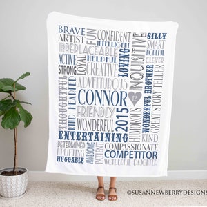 May include: White baby blanket with blue and gray text. The text includes the name "Connor" and the year "2015". The blanket also features a heart with the number "12" inside.