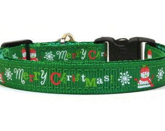May include: Green Christmas-themed cat collar with a black buckle and white text that reads "Merry Christmas!" with red and green accents. The collar features a repeating pattern of snowmen and snowflakes.