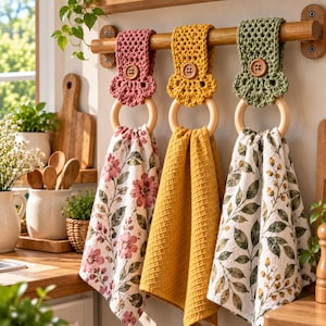 Op de afbeelding: Drie hangende keukendoeken aan een houten stang. Elke handdoek heeft een gehaakte bovenkant in roze, geel of groen met een houten knoop, bevestigd aan een houten ring. De doeken hebben bloemmotieven, een wafelstructuur en bladpatronen.