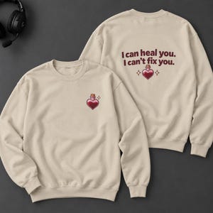 May include: A beige crewneck sweatshirt featuring a small heart-shaped potion bottle graphic on the front left chest. The back displays the text "I can heal you. I can't fix you." in dark red, accompanied by a heart-shaped potion bottle and star icons.