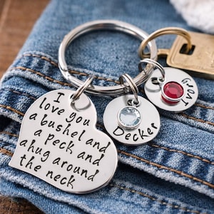 May include: A silver keychain with multiple charms. One charm is heart-shaped with the words "I love you a bushel and a peck and a hug around the neck." Other charms include names and gemstones. The keychain rests on denim.