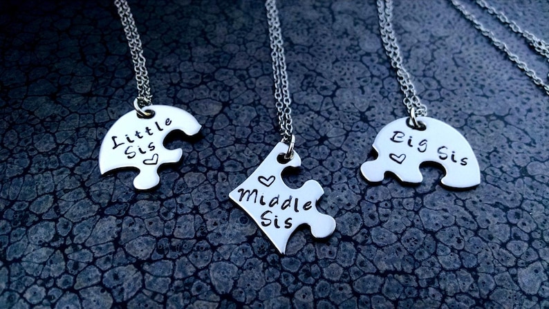 May include: Three silver puzzle piece necklaces. Each piece is engraved with "Little Sis", "Middle Sis", and "Big Sis" with a heart symbol. The necklaces hang on a silver chain.