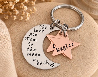 May include: A silver-toned metal keychain featuring a crescent moon charm engraved with the text We love you Mom to the moon & back and a small heart. Two copper-toned star charms are attached, one of which is engraved with the name Kaylee.