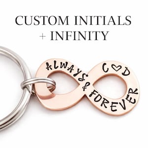 May include: A rose gold keychain featuring an infinity symbol with the words "ALWAYS & FOREVER" and "GOD" with a heart symbol. The keychain also has the text "CUSTOM INITIALS + INFINITY" in black. The keychain is attached to a silver metal ring.