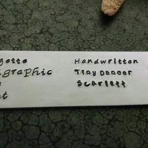 May include: A white metal tag with black lettering. The text reads "Bridgette Calligraphic Dots script" and "Handwritten Tiny Dancer Scarlett".