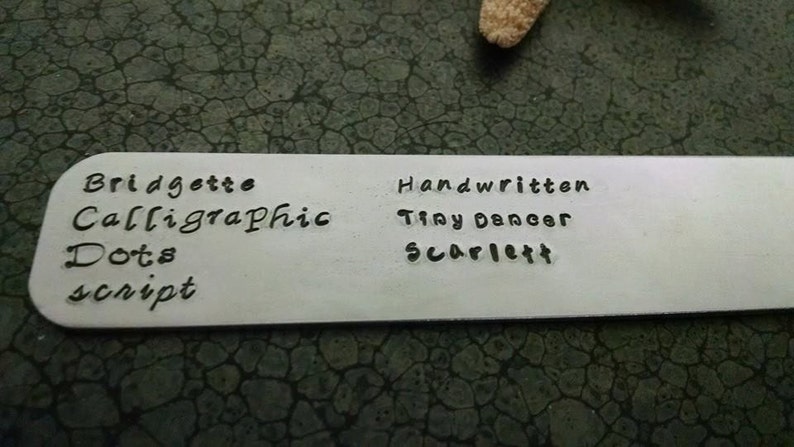 May include: A white metal tag with black lettering. The text reads "Bridgette Calligraphic Dots script" and "Handwritten Tiny Dancer Scarlett".