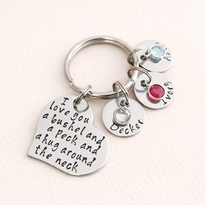 May include: A silver-colored keychain with a heart-shaped charm engraved with the phrase "I love you a bushel and a peck and a hug around the neck." It includes round charms with names and gemstones.