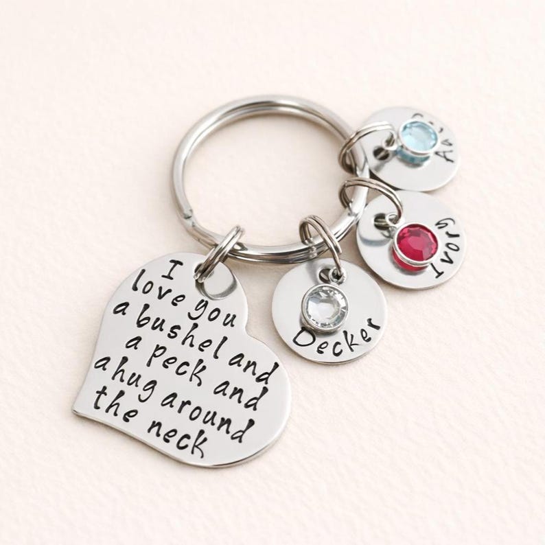 May include: A silver-colored keychain with a heart-shaped charm engraved with the phrase "I love you a bushel and a peck and a hug around the neck." It includes round charms with names and gemstones.