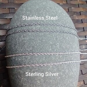May include: Close-up of a gray stone with two silver chains and two stainless steel chains. The text "Stainless Steel" and "Sterling Silver" is printed on the stone.