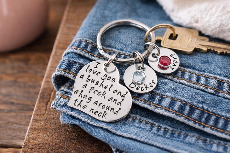 May include: A silver keychain with multiple charms. One charm is heart-shaped with the words "I love you a bushel and a peck and a hug around the neck." Other charms include names and gemstones. The keychain rests on denim.