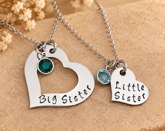 May include: Two silver-toned necklaces featuring heart-shaped pendants. One large open heart pendant is engraved with the text Big Sister and includes a dark green circular stone. A smaller solid heart pendant is engraved with Little Sister and a light blue stone.