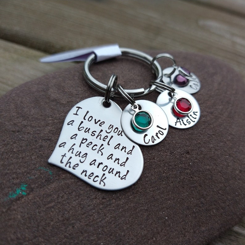 May include: A silver keychain with a heart-shaped charm engraved with the phrase "I love you a bushel and a peck and a hug around the neck." Additional charms include round pendants with names and colored gemstones.