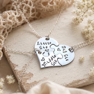 May include: Silver necklaces with a heart-shaped pendant split into three puzzle pieces. Each piece is engraved with "Little Sis," "Middle Sis," and "Big Sis," with heart accents. The necklaces are displayed on a wooden surface.