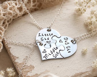 May include: Silver necklaces with a heart-shaped pendant split into three puzzle pieces. Each piece is engraved with "Little Sis," "Middle Sis," and "Big Sis," with heart accents. The necklaces are displayed on a wooden surface.