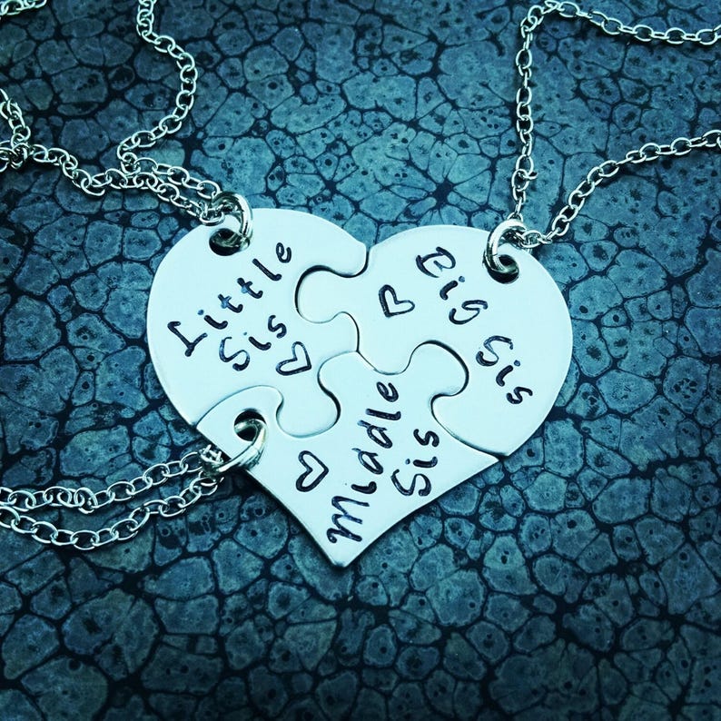 May include: A silver necklace featuring a heart-shaped pendant made of four puzzle pieces. Each piece is engraved with "Little Sis", "Big Sis", and "Middle Sis", along with heart symbols. The necklace has a delicate chain.