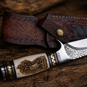 Puede incluir: Un cuchillo de hoja fija con una hoja de metal plateado que presenta una textura martillada cerca del lomo. El mango es de color crema con una imagen grabada de una cabeza de ciervo y detalles metálicos. Incluye una funda de cuero marrón.