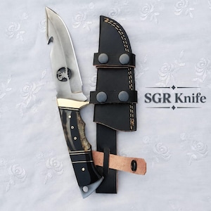 Puede incluir: Un cuchillo de hoja fija con gancho, una hoja plateada con un grabado circular de cabeza de ciervo y un mango de cuerno oscuro con detalles en latón. Incluye una funda de cuero negro con cierres a presión y el texto SGR Knife.