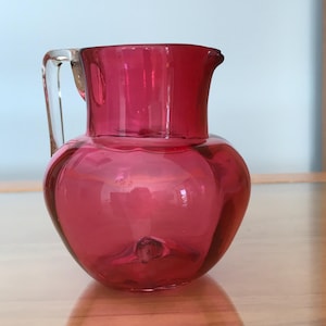 Puede incluir: Una pequeña jarra de vidrio bulbosa en un vibrante color magenta translúcido. El recipiente presenta un cuerpo ancho y redondeado, un cuello corto, un pequeño pico vertedor y un asa de vidrio transparente y curva unida al costado.