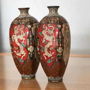 May include: A pair of matching vases featuring a vibrant red central panel decorated with a white dragon motif. The lower section has a textured brown finish with gold flecks, while the neck and upper body display intricate geometric patterns.