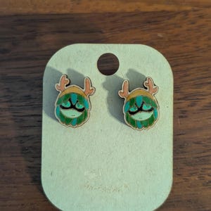 May include: A pair of wooden stud earrings featuring a whimsical green and teal creature design with antlers. The earrings are mounted on a light brown card with a small hole at the top. The card is on a wooden surface.