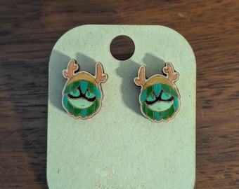 May include: A pair of wooden stud earrings featuring a whimsical green and teal creature design with antlers. The earrings are mounted on a light brown card with a small hole at the top. The card is on a wooden surface.