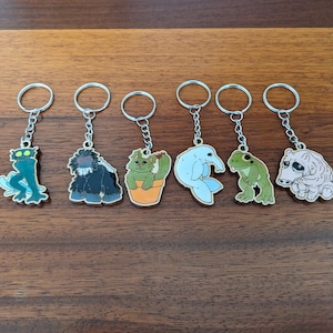 May include: Seven keychains with different cartoon characters. The characters include a green monster, a black gorilla, a green plant, a blue dolphin, a green frog, and a pink worm. The keychains are all silver.
