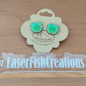 May include: A pair of green and blue wooden earrings with a cartoon face design. The earrings are displayed on a wooden tag with the text "LaserFish Creations" and an email address. The tag is on a wooden surface. The text "@LaserFishCreations" is on a clear acrylic sign.