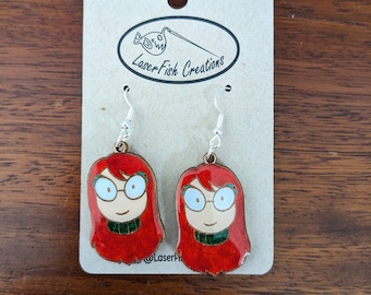 May include: A pair of earrings featuring a cartoon illustration of a person with red hair, glasses, and a green shirt. The earrings are on a card that says "LaserFish Creations".
