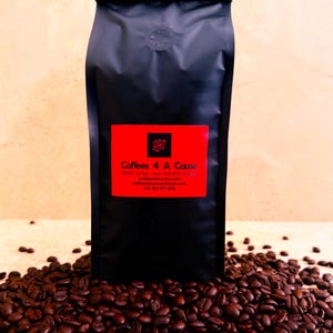 May include: A matte black coffee bag with a rectangular red label featuring the text Coffees 4 A Cause, a coffee bean icon, and contact details. The bag is surrounded by a pile of dark roasted coffee beans on a light surface.