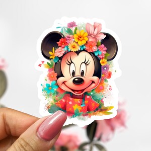 May include: A colorful sticker featuring Minnie Mouse wearing a floral crown and a red dress with a floral pattern. The sticker has a white border and is held by a hand with a pink nail polish.