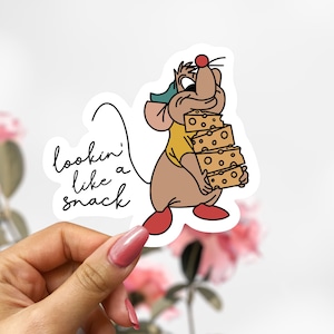 May include: A die-cut sticker featuring a cartoon mouse carrying a stack of cheese. The mouse is wearing a yellow shirt, a green hat, and red shoes. The sticker has the text "lookin' like a snack" in a handwritten font.