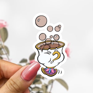 May include: A white teacup with a smiling face and a gold handle, with brown liquid and bubbles inside. The teacup is surrounded by pink and white flowers.
