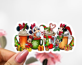 May include: A sticker featuring a row of five Christmas-themed coffee cups with Mickey Mouse and Minnie Mouse characters. Each cup has a different character and a festive design with red, green, and white colours. The cups are decorated with snowflakes, candy canes, and Christmas trees.