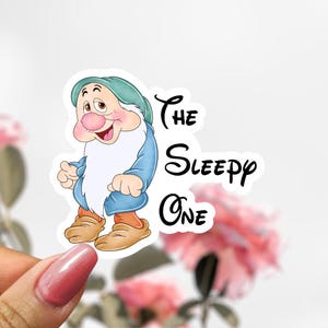 May include: A sticker featuring the character Sleepy from Snow White. Sleepy is depicted with a blue outfit, a green hat, and a long white beard. The text "The Sleepy One" is written in black script.