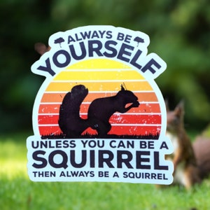 May include: A sticker with a retro sunset design and a black squirrel silhouette. The text reads "ALWAYS BE YOURSELF UNLESS YOU CAN BE A SQUIRREL THEN ALWAYS BE A SQUIRREL" in a playful font.