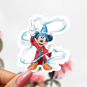 May include: A sticker featuring Mickey Mouse in a sorcerer's outfit. Mickey is wearing a blue hat with stars and a red robe. Blue magic swirls around him as he points upwards. The sticker has a white border.