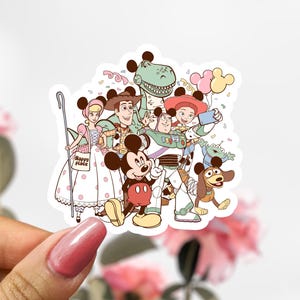 Peut inclure: Un autocollant représentant un groupe de personnages Disney, dont Mickey Mouse, Buzz l'Éclair et Woody. Les personnages sourient et posent ensemble, avec des confettis et des ballons en arrière-plan. L'autocollant a une bordure blanche.