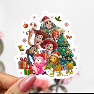 Peut inclure: Un autocollant coloré représentant les personnages de Toy Story célébrant Noël. Woody, Buzz l'Éclair, Jessie, Hamm et Zigzag sont représentés avec un sapin de Noël, des cadeaux et des flocons de neige. L'autocollant est tenu par une personne.